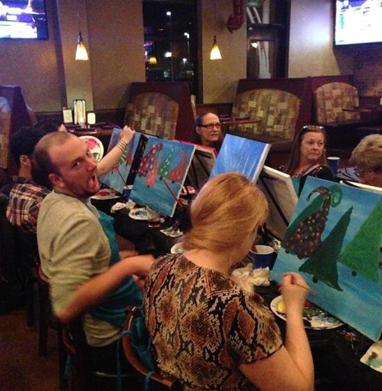 Guests at a restaurant table reacting playfully to their finished paintings during a Painting Party Events class.