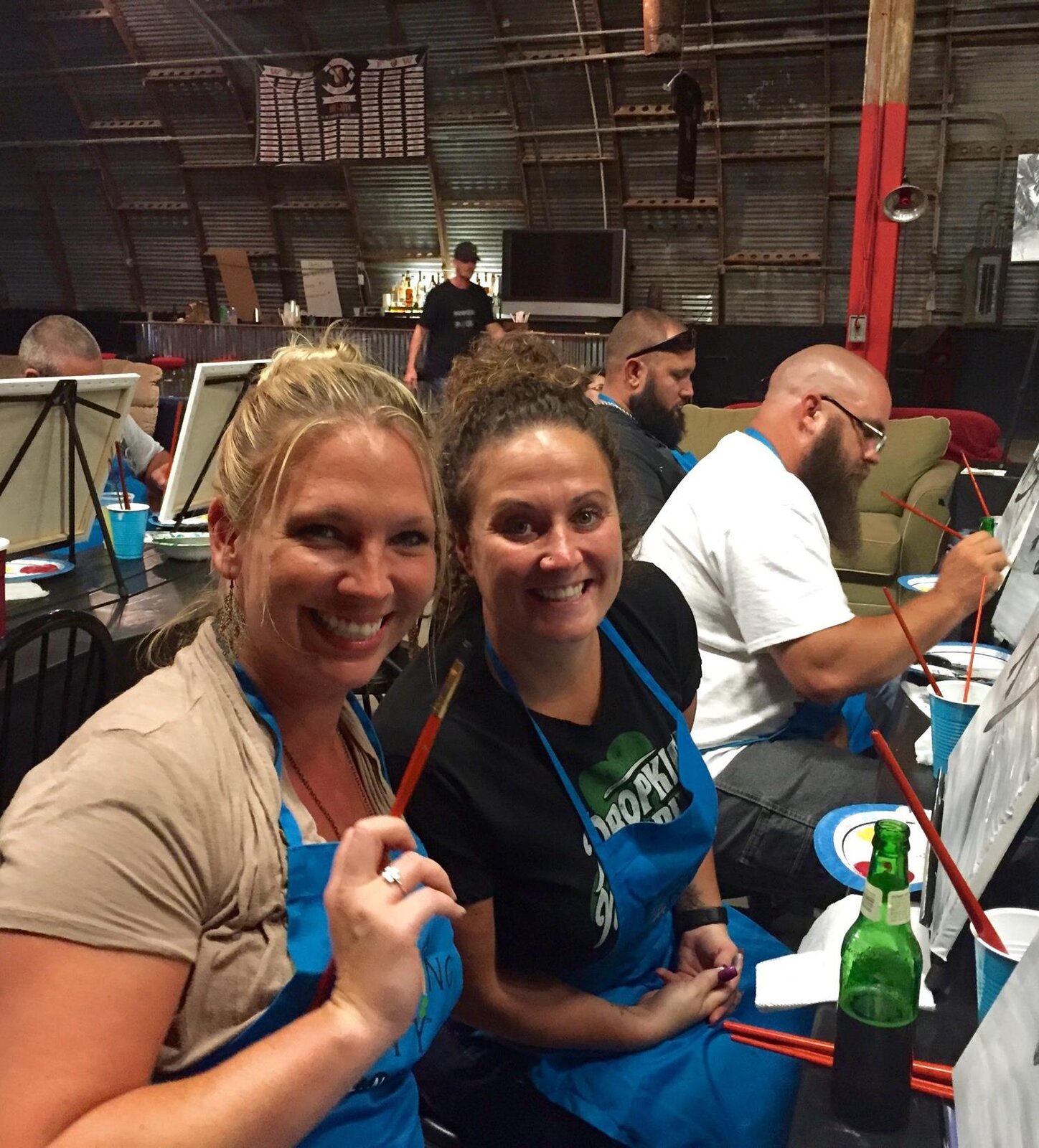Two guests in blue Painting Party Events aprons smiling and holding paintbrushes at an event.
